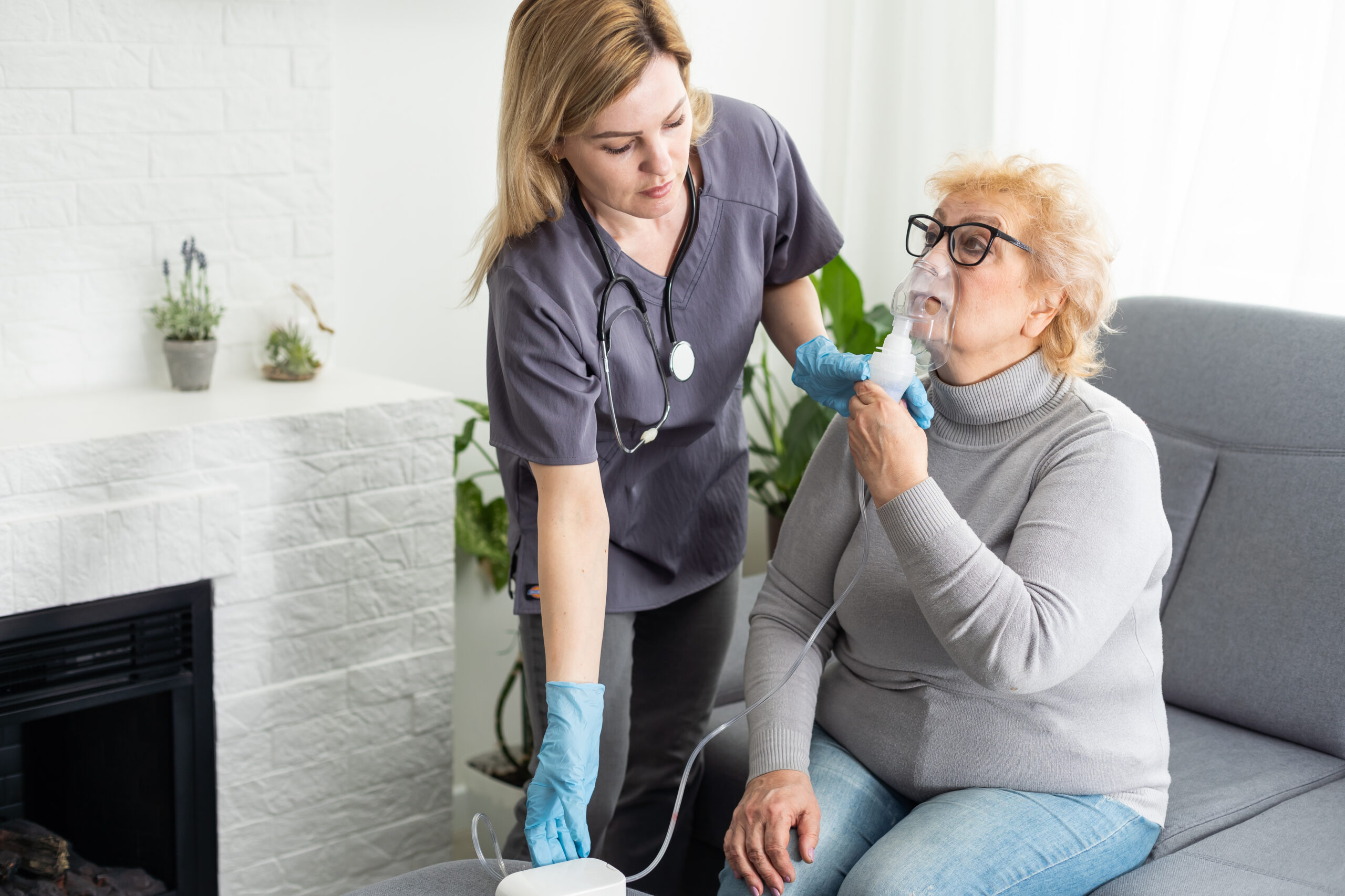 Young high-skilled pleasant woman doctor putting on nebulizer mask on face of her elderly female patient to make inhalation when visitng him at home. Flu, cold and cough treatment