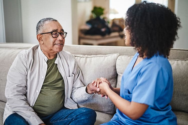 Nurse assistant holds hand of senior man on sofa at assisted living facility